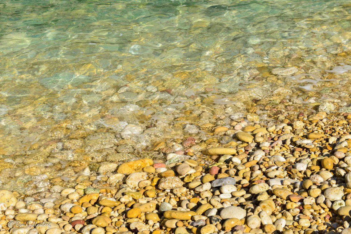 Klart turkost vatten och små gyllene stenar på stranden.