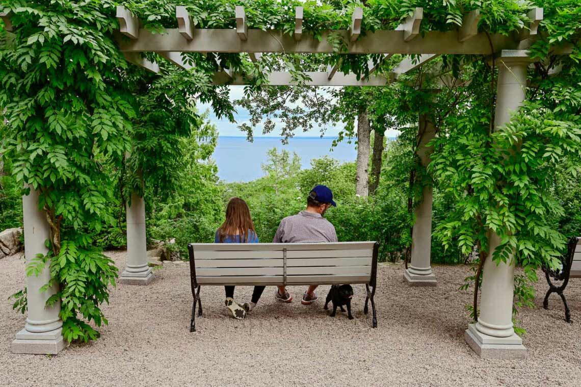 två människor och en hund sitter på en bänk och tittar ut över havet under en pergola
